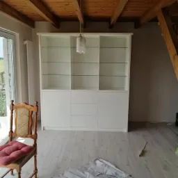 Biała szafa z przeszklonymi półkami i trzema szufladami, widoczna w przestronnym pokoju z drewnianym sufitem i schodami, na podłodze leżą szare spodnie i ołówek.