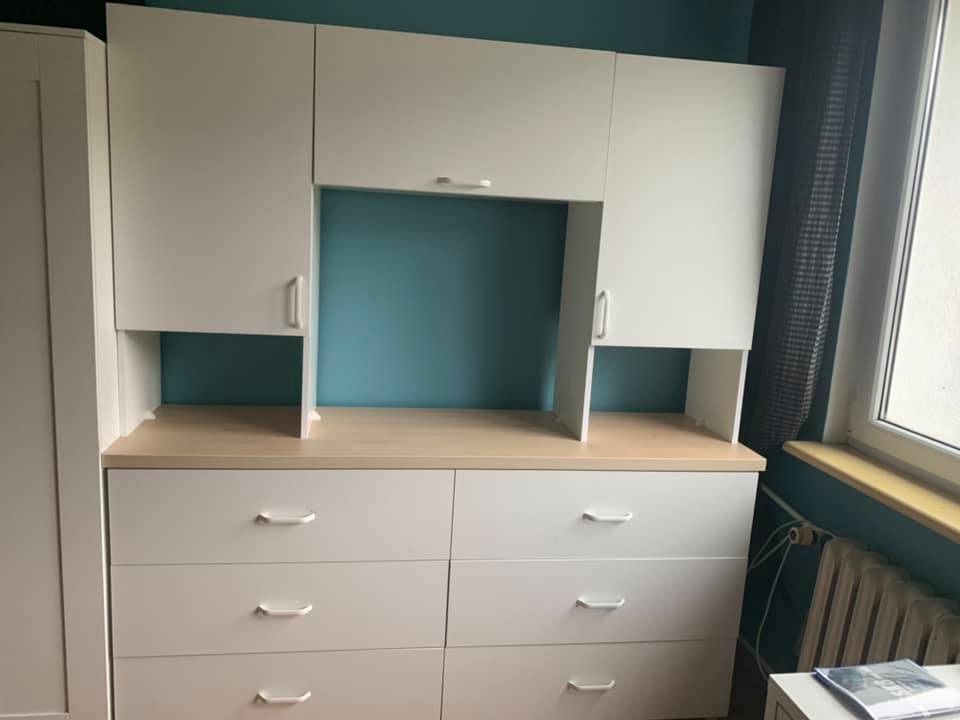 Biała szafa z szufladami i szafkami wiszącymi na tle turkusowej ściany, z widocznym fragmentem grzejnika pod oknem.