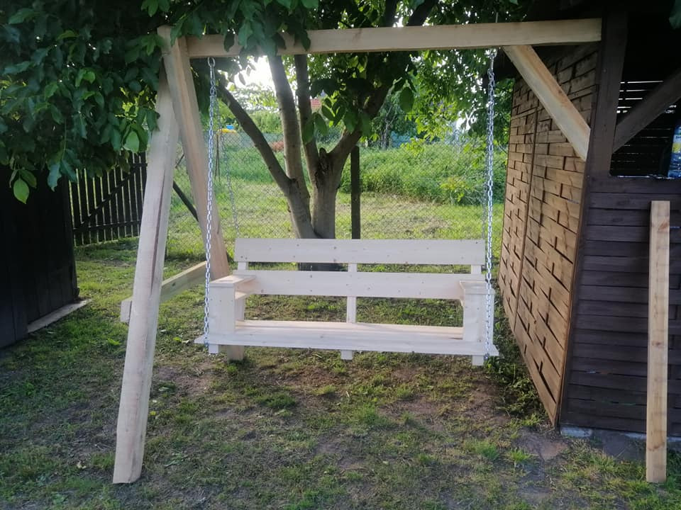 Surowa, drewniana huśtawka ogrodowa zawieszona na łańcuchach, w tle zielony ogród i drewniana altana, widoczne niedoskonałości obróbki drewna.