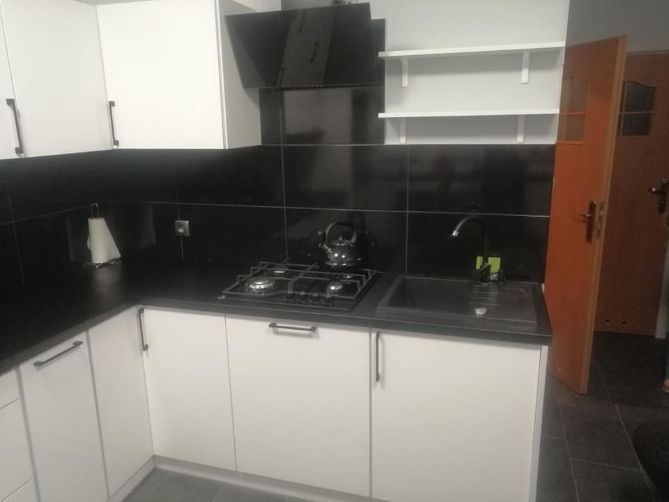 Nowoczesna kuchnia z białymi szafkami, czarnym blatem i płytkami, widoczny zlew, kuchenka gazowa i półki na ścianie.