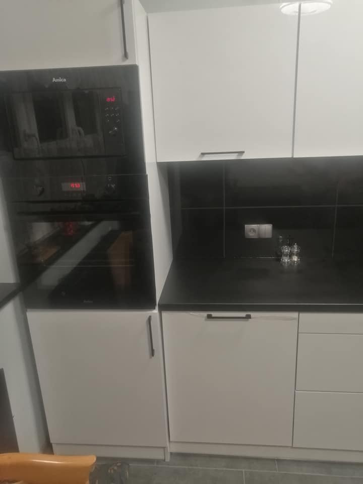 Nowoczesna zabudowa kuchenna z białymi frontami, czarnym blatem i sprzętem AGD marki Amica, widok z niskiej perspektywy.