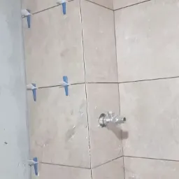 Ściana łazienki w trakcie układania beżowych płytek, widoczne niebieskie klipsy poziomujące i wystające rury instalacji wodnej.