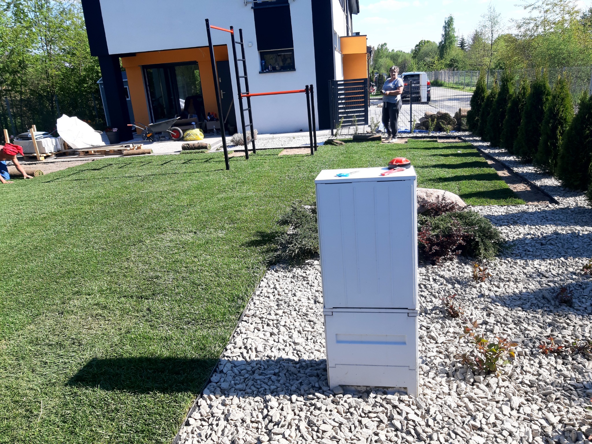 Układanie trawy z rolki w ogrodzie, widoczna biała skrzynia narzędziowa, osoba pracująca przy rozwijaniu trawy oraz kobieta obserwująca postęp prac, nowoczesny dom w tle.