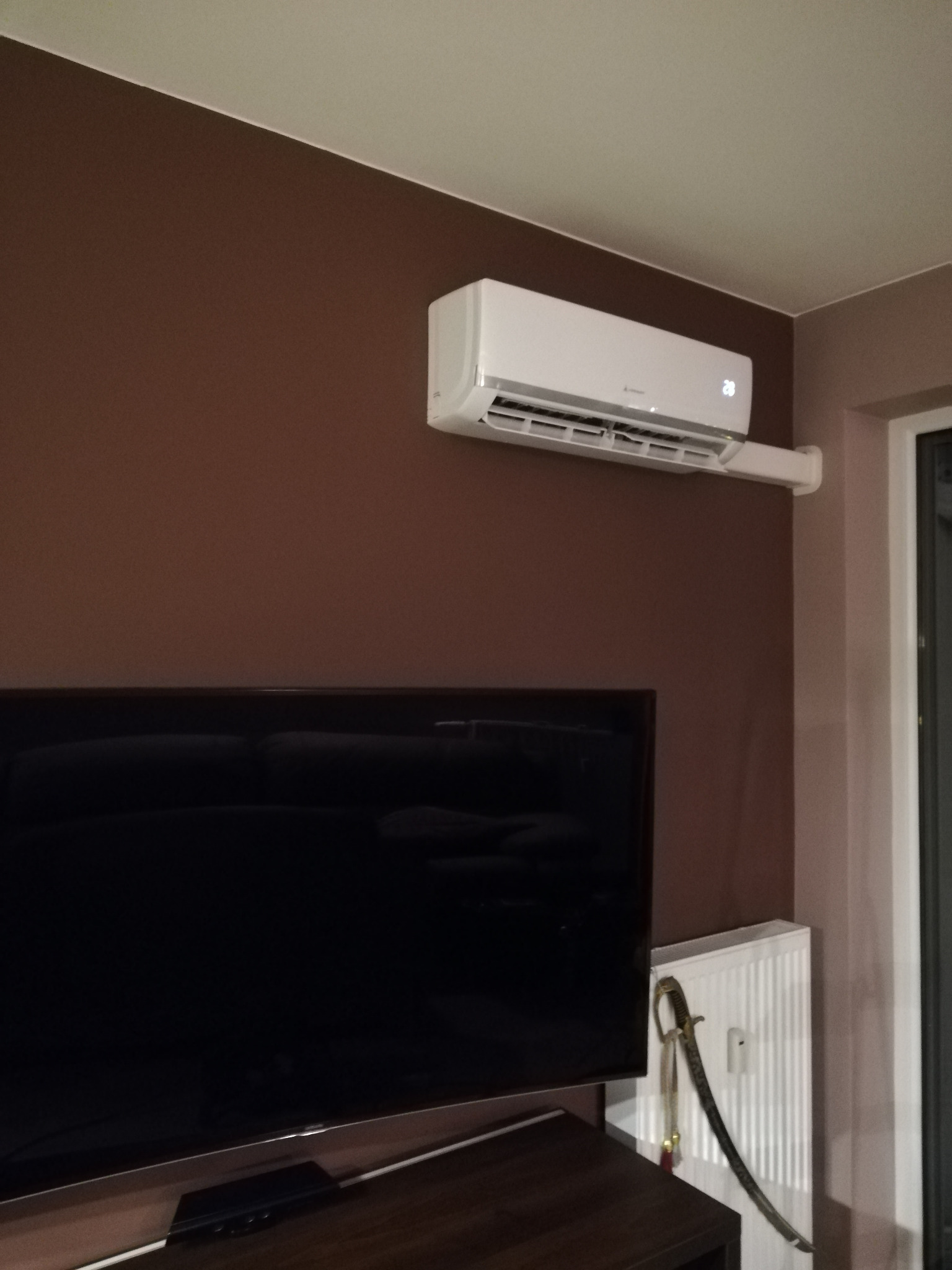 Biały klimatyzator na brązowej ścianie nad telewizorem, szabla dekoracyjna oparta o grzejnik w rogu pokoju.