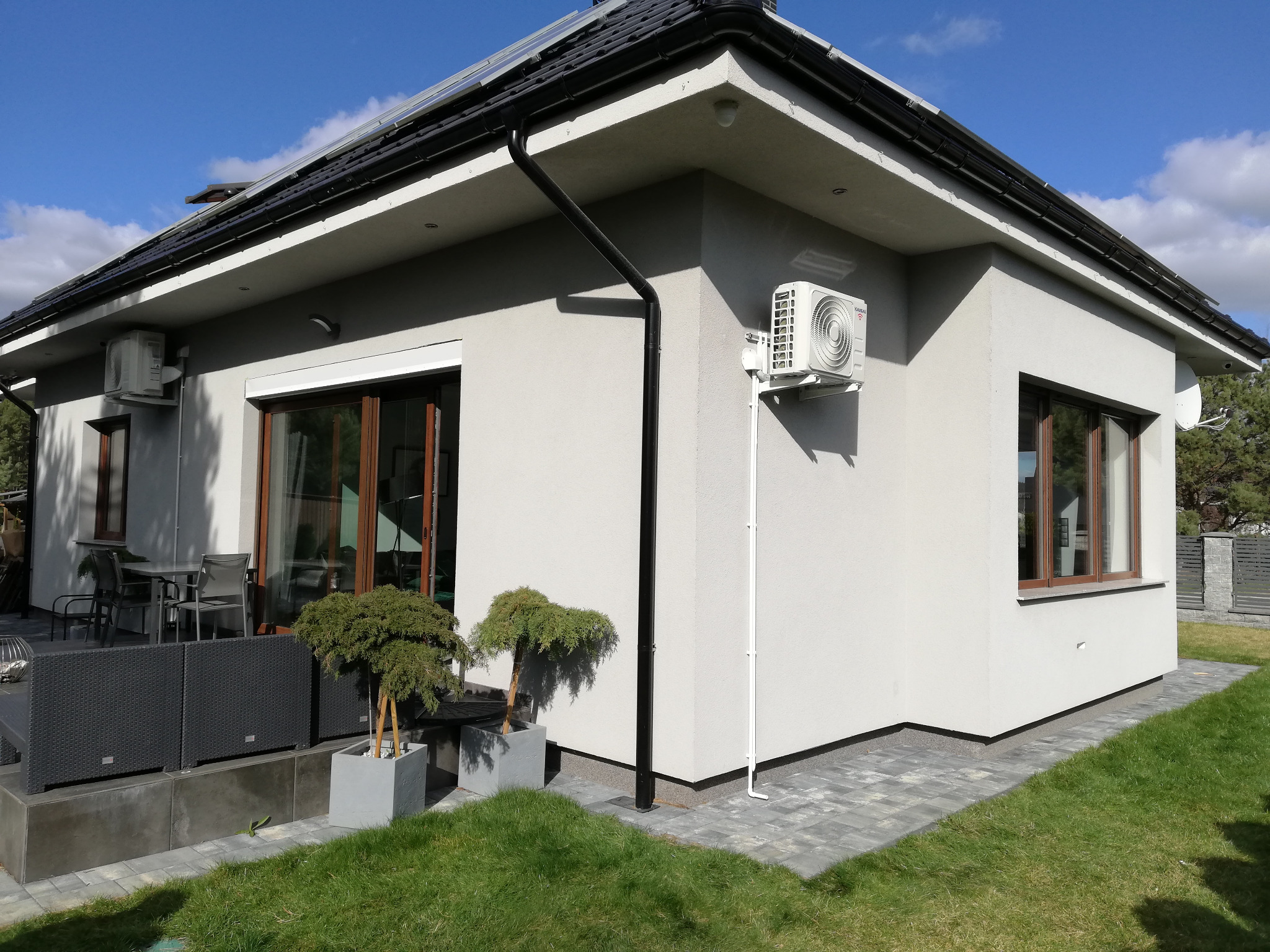 Zewnętrzna jednostka klimatyzatora marki KAISAI zamontowana na jasnej elewacji domu jednorodzinnego, widoczna rynna i instalacja odprowadzająca skropliny, w tle fragment tarasu z meblami ogrodowymi...