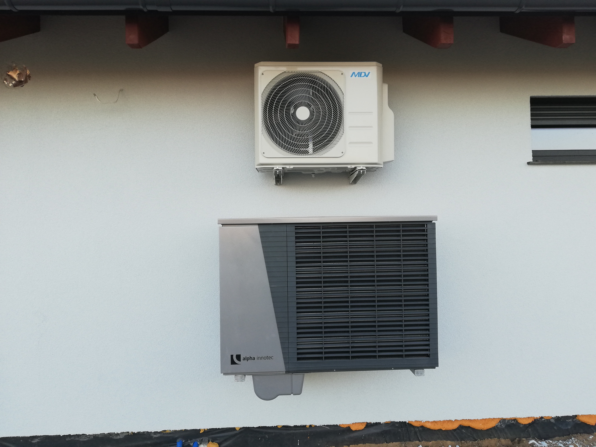 Zewnętrzne jednostki klimatyzacji MDV i pompy ciepła Alpha Innotec zamontowane na elewacji budynku z widocznymi elementami izolacji i orynnowania.