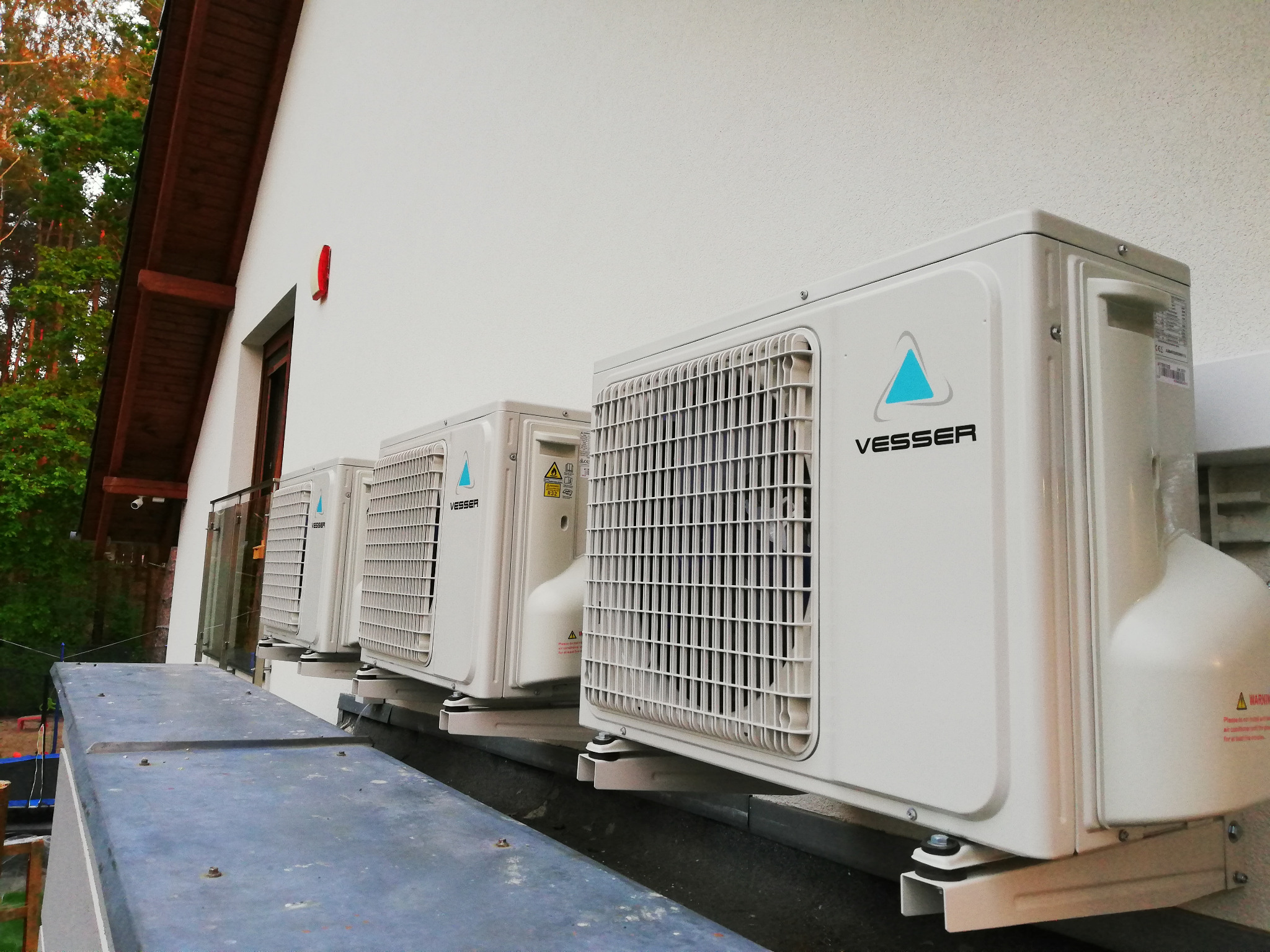 Trzy zewnętrzne jednostki klimatyzacji Vesser zamontowane na ścianie budynku z widocznymi wspornikami montażowymi i tabliczkami ostrzegawczymi.