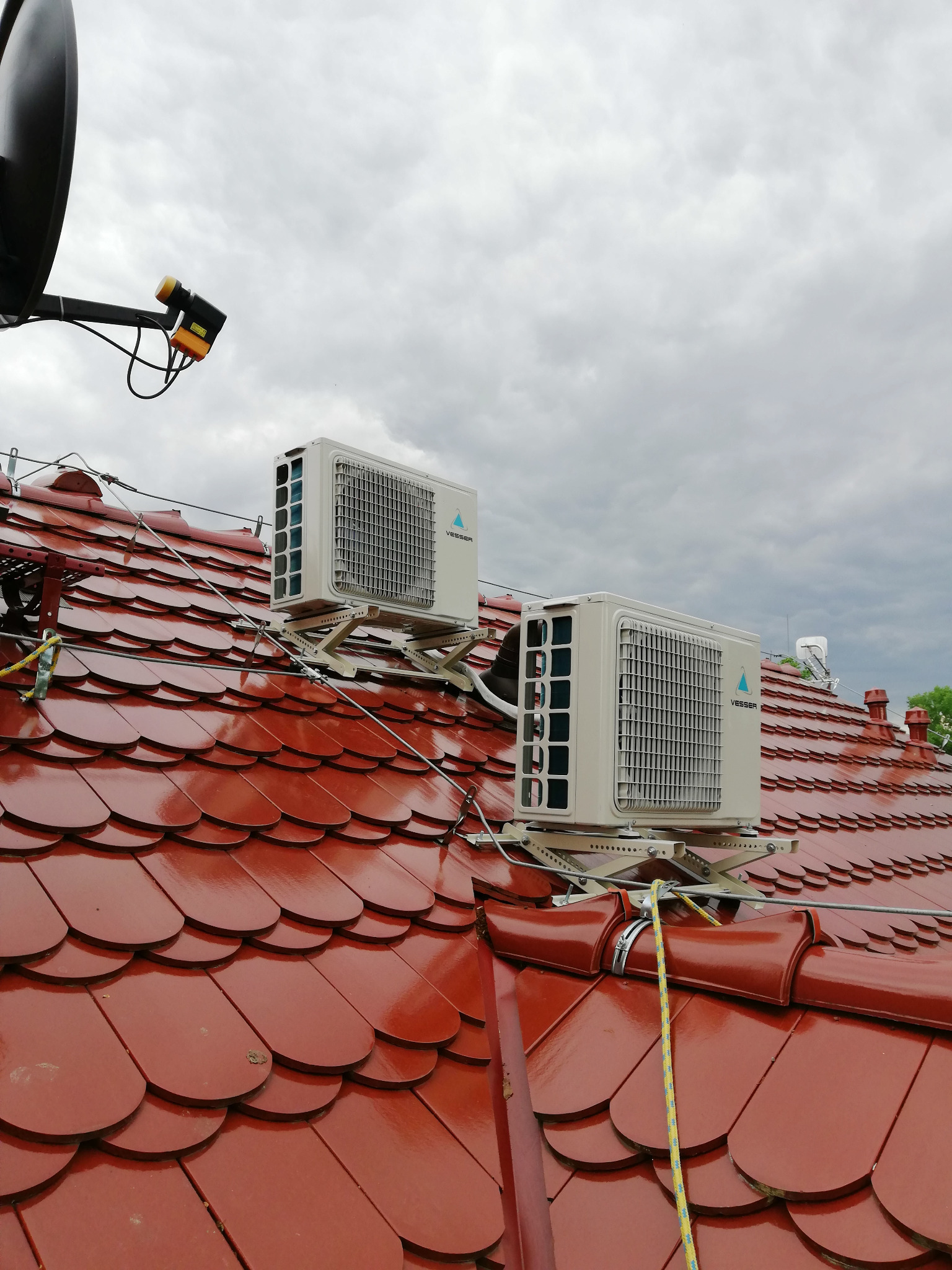 Dwa zewnętrzne moduły klimatyzacyjne Vesser zamontowane na specjalnych wspornikach na błyszczącym, czerwonym dachu z dachówki ceramicznej, widoczne liny zabezpieczające.