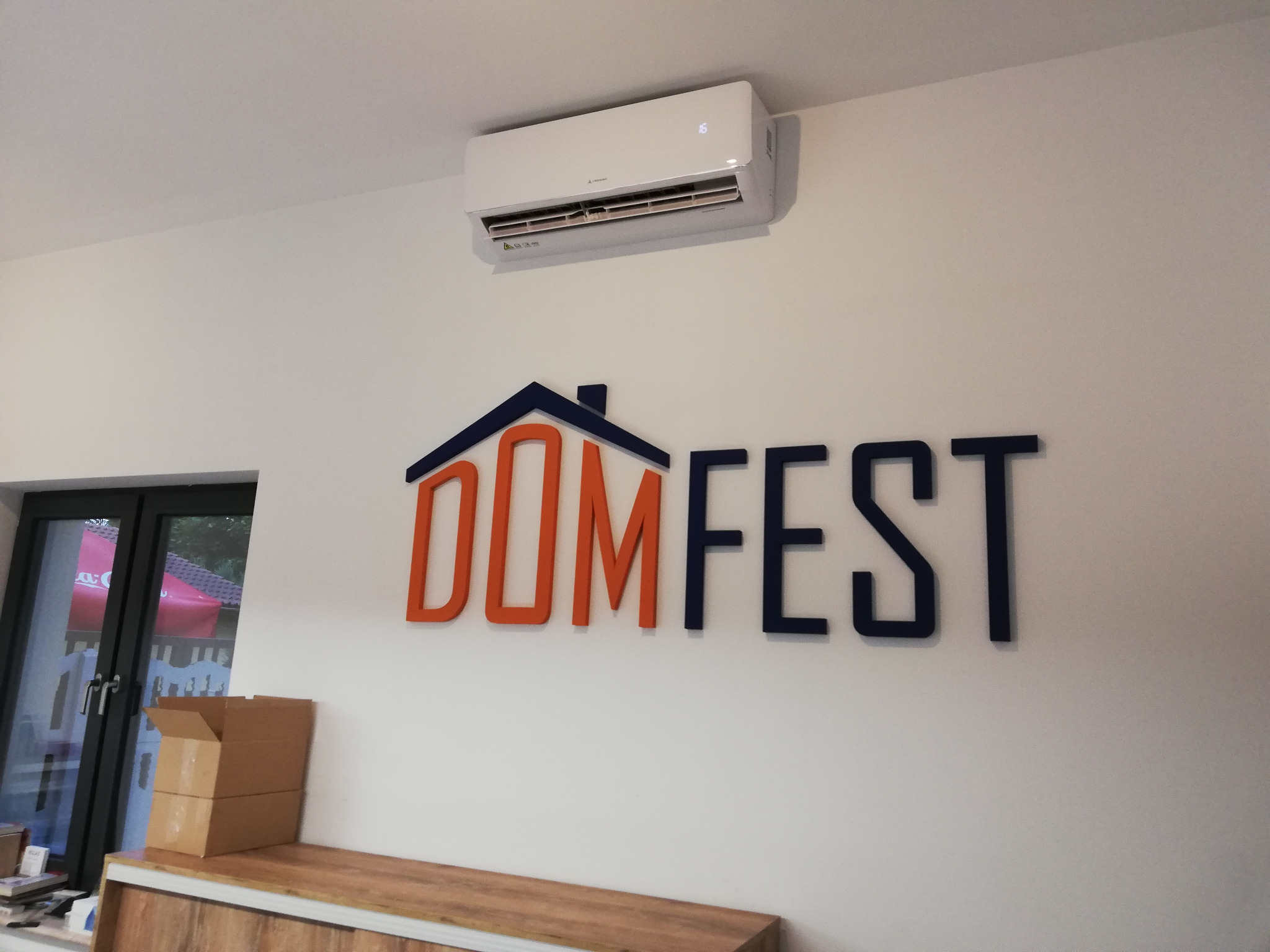 Biała jednostka klimatyzacyjna zamontowana na ścianie nad napisem 'DOM FEST', widok z dołu, w tle okno.
