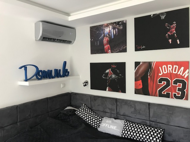 Szary klimatyzator ścienny w pokoju z dekoracjami sportowymi, niebieski napis 'Domuvik' na białej półce, cztery obrazy z Michaelem Jordanem na ścianie, szara sofa narożna z poduszkami.