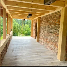 Zadaszony taras z drewnianą konstrukcją, ceglaną ścianą i podłogą z płytek imitujących drewno, widok na ogród.