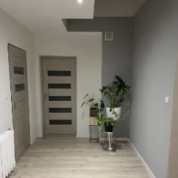 Jasne panele podłogowe w korytarzu z dwiema szarymi drzwiami z przeszkleniami, szara ściana z wentylacją, biała ściana, rośliny doniczkowe na metalowych stojakach.