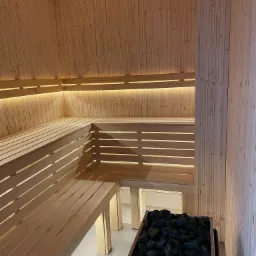 Wnętrze wykończonej drewnem jasnej kabiny z ławkami i podświetleniem LED, z piecem na czarne kamienie w dolnym rogu.