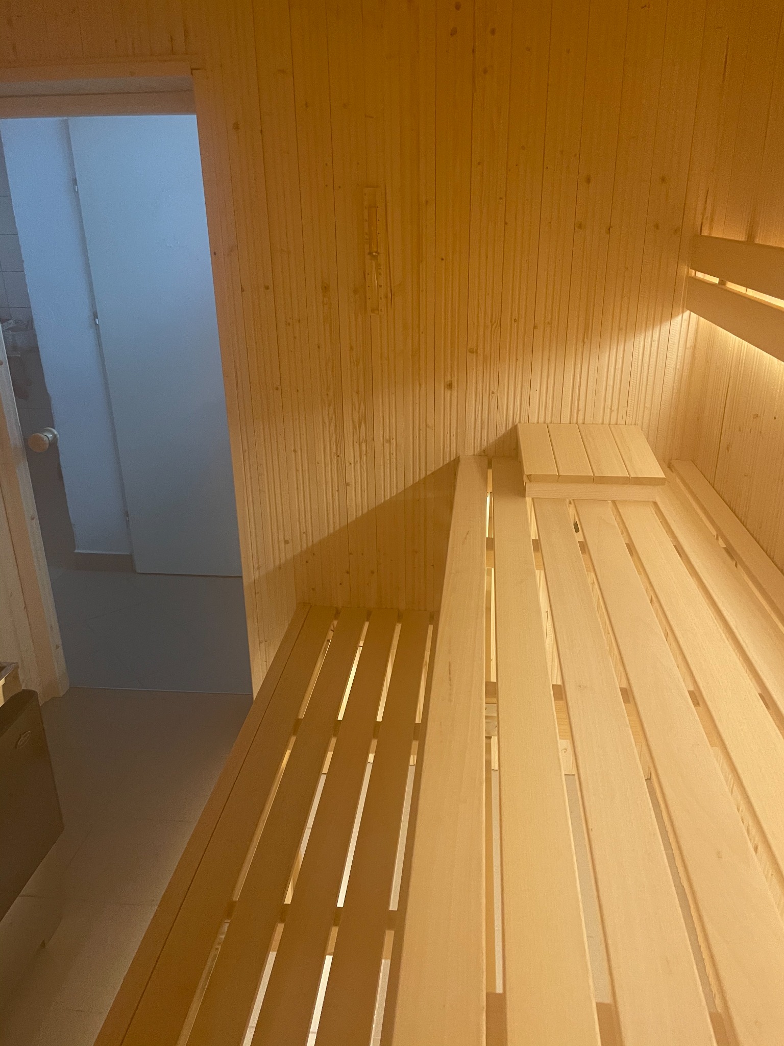 Wnętrze drewnianej kabiny z ławkami, oświetlone ciepłym światłem, z widocznym wejściem do pomieszczenia z jasnymi płytkami.