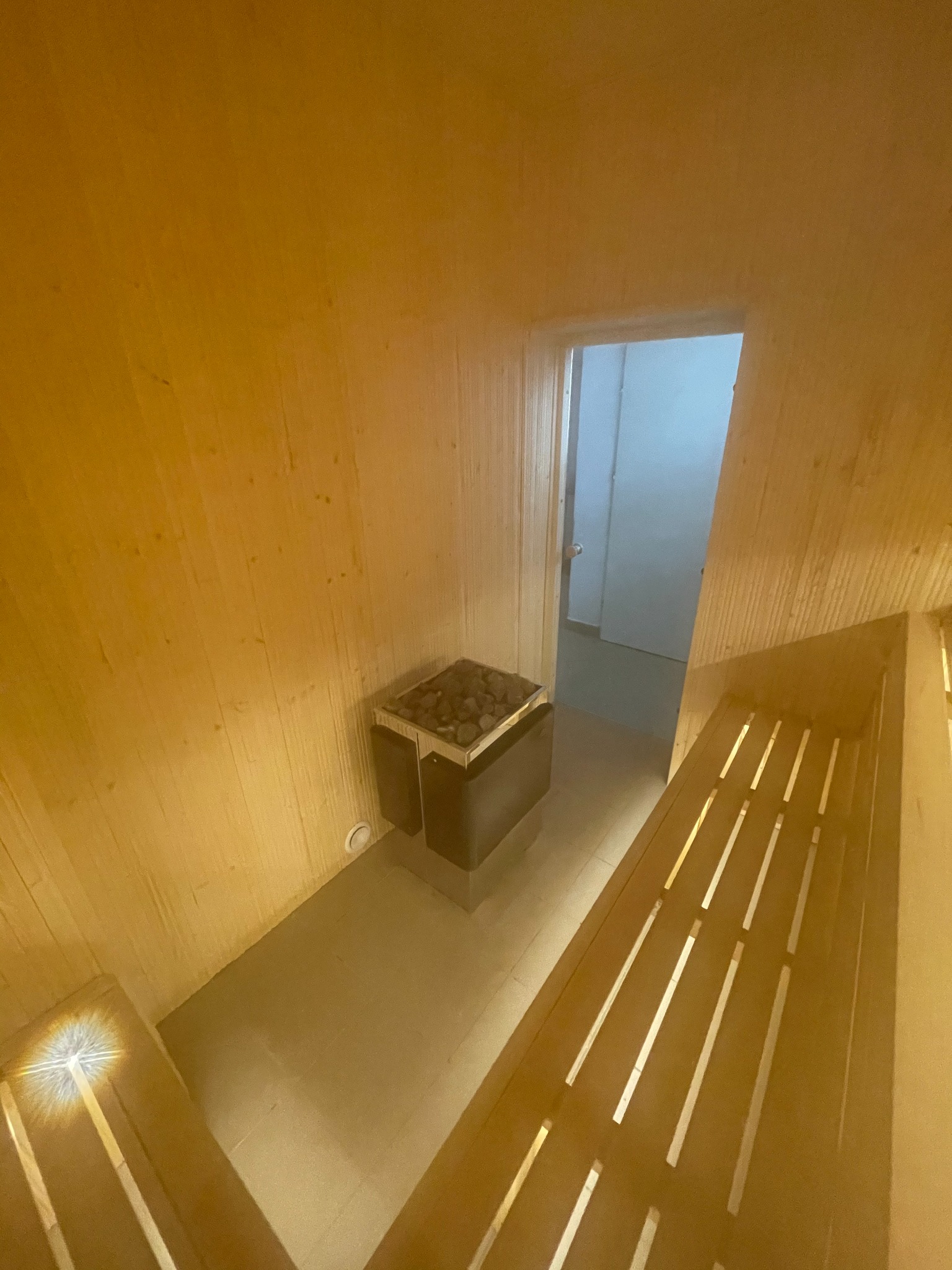 Wnętrze wykończonej drewnem kabiny z piecem na kamienie i drewnianymi ławkami z podświetleniem LED w szczelinach, widok na drzwi wyjściowe.