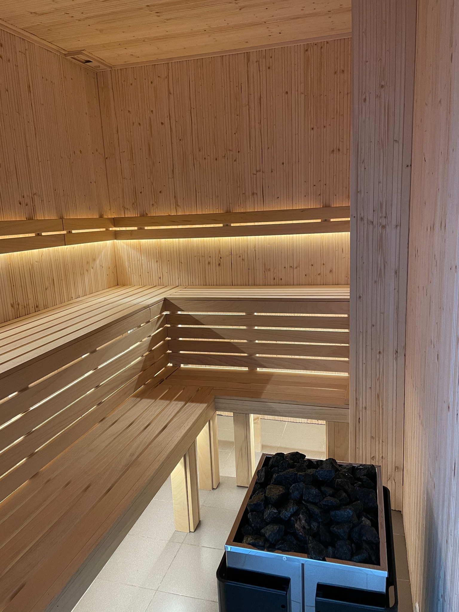 Wnętrze wykończonej drewnem jasnej kabiny z ławkami i podświetleniem LED, z piecem na czarne kamienie w dolnym rogu.