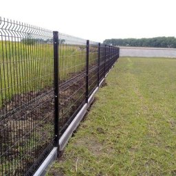 FIRMA BUDOWLANA GRZEGORZ ZAJĄC - Długi, czarny, metalowy system ogrodzeniowy z panelami na betonowej podmurówce, oddzielający trawnik od świeżo przekopanej ziemi, z widokiem na odległe drzewa i betonowy mur.