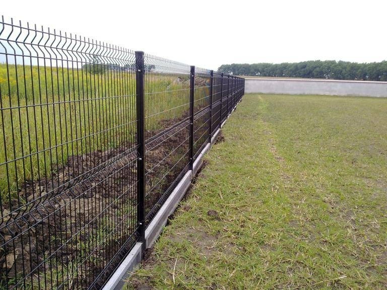 Długi, czarny, metalowy system ogrodzeniowy z panelami na betonowej podmurówce, oddzielający trawnik od świeżo przekopanej ziemi, z widokiem na odległe drzewa i betonowy mur.