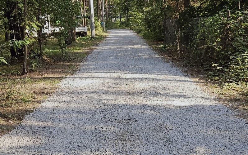 Świeżo utwardzona droga żwirowa biegnąca przez las, widoczny fragment zaparkowanego pojazdu w tle.