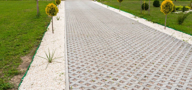 Długa, szara droga z ażurowych betonowych płyt wypełnionych żwirem, obrzeża wyłożone białym kamieniem, trawnik z niskimi, kulistymi drzewkami po bokach.