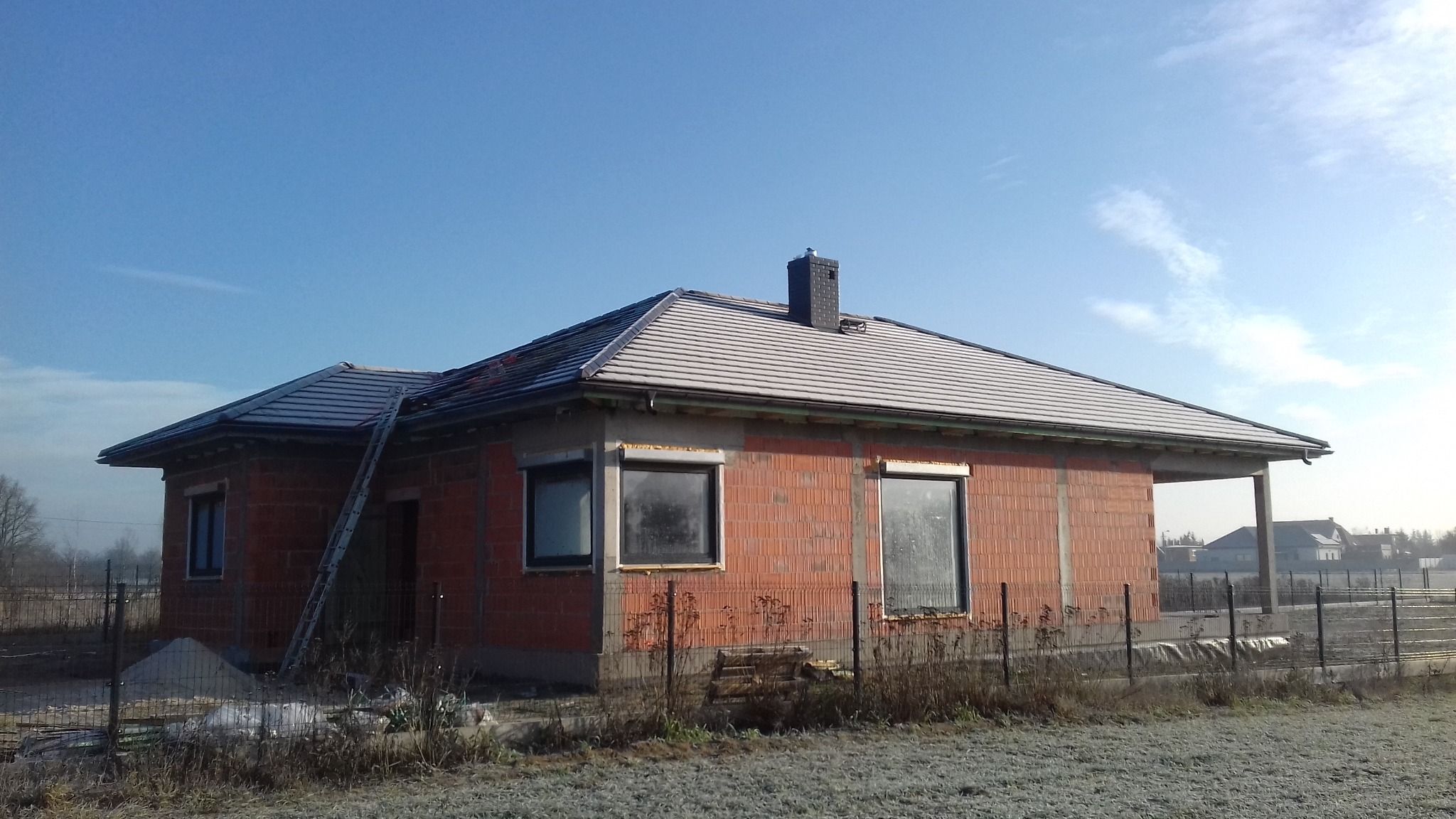 Surowy budynek jednorodzinny z częściowo pokrytym dachem dachówką, widoczna drabina oparta o ścianę, stan surowy otwarty.