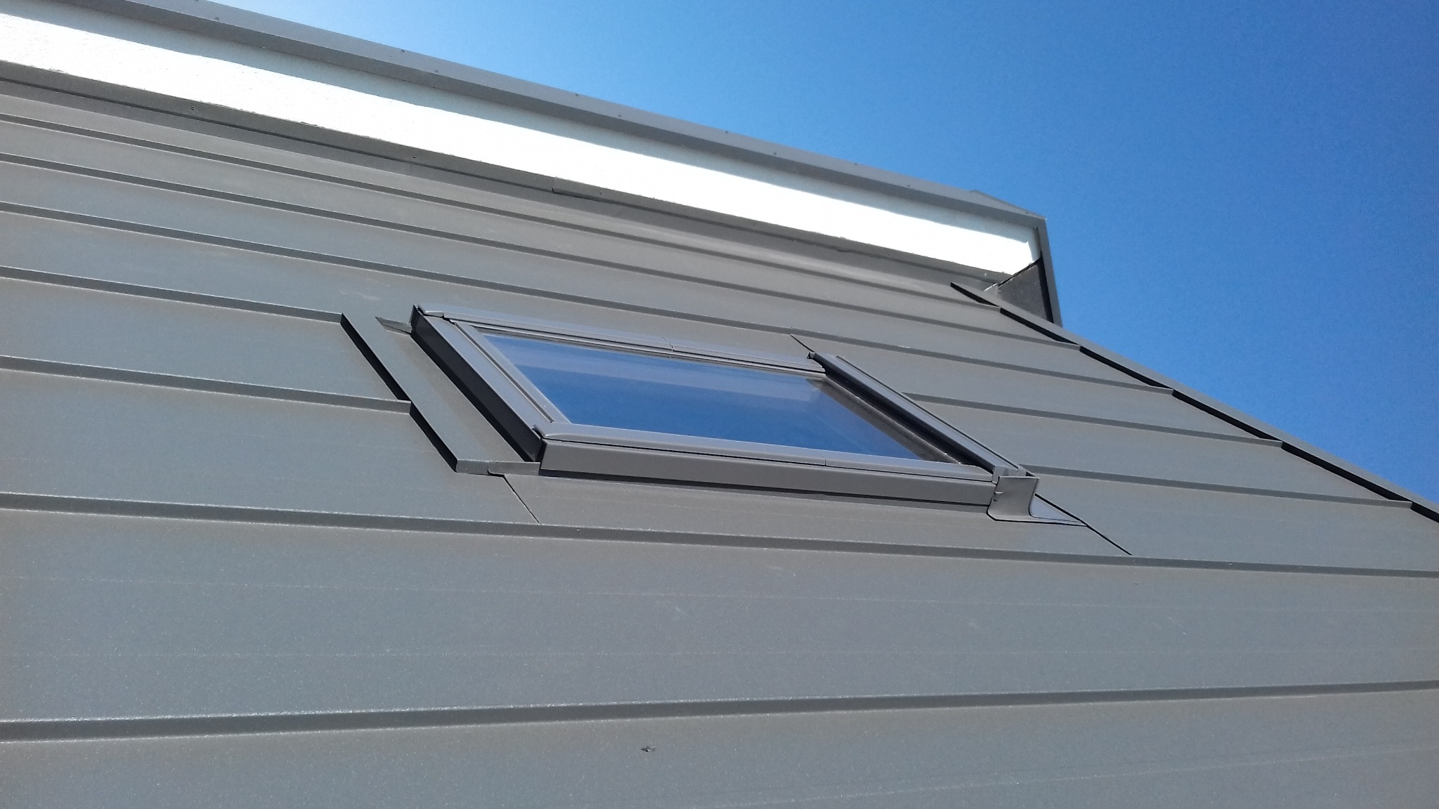 Szary dach z blachy na rąbek stojący z oknem dachowym, widok z dołu na tle błękitnego nieba.