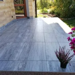 Zadaszony taras z szarymi płytkami imitującymi drewno, kwiaty w donicach, widok na ogród.