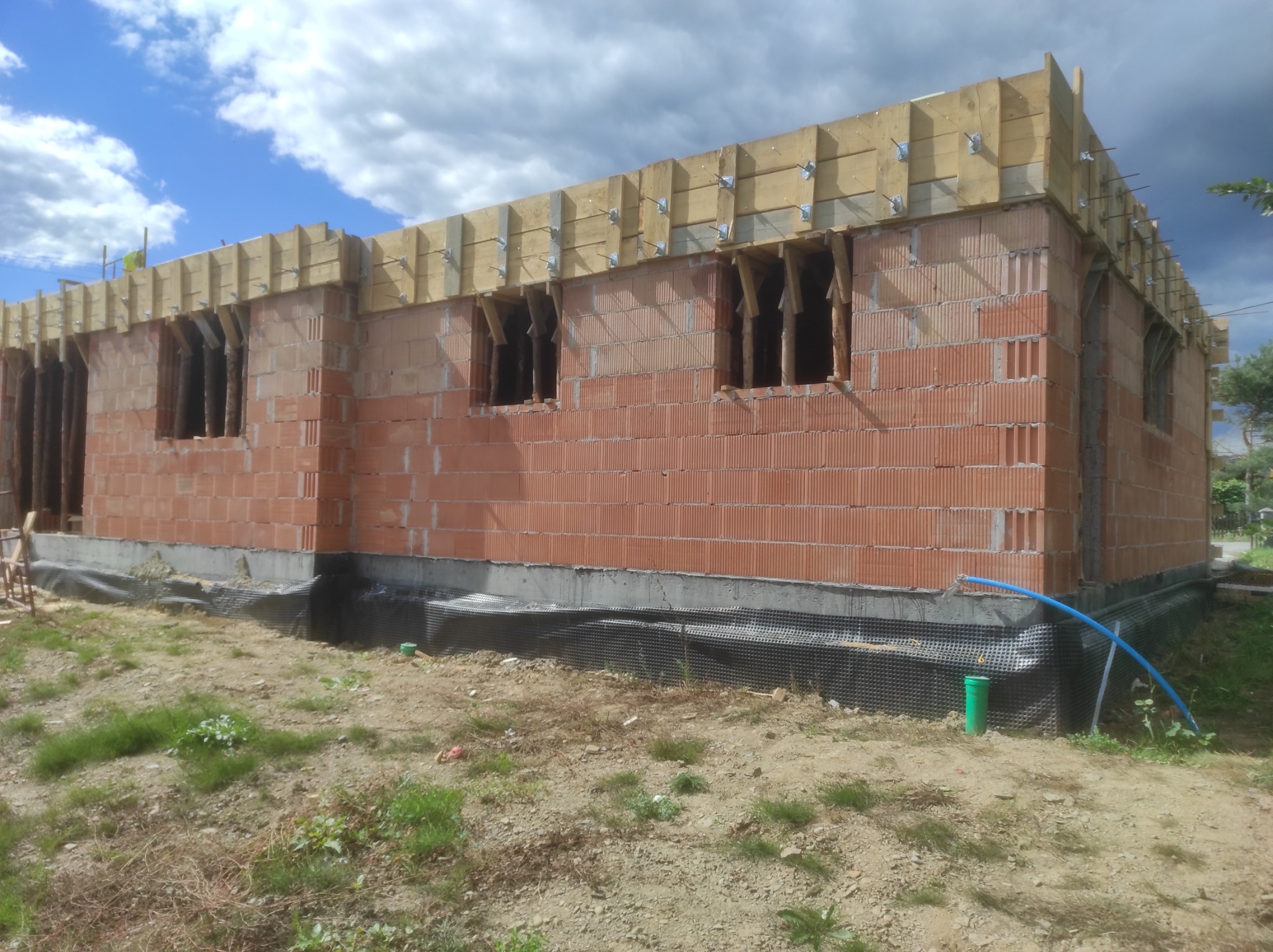 Wznoszenie ścian domu z czerwonej cegły, widoczne drewniane szalunki na górze, fundament zabezpieczony folią kubełkową, teren wokół w stanie surowym.