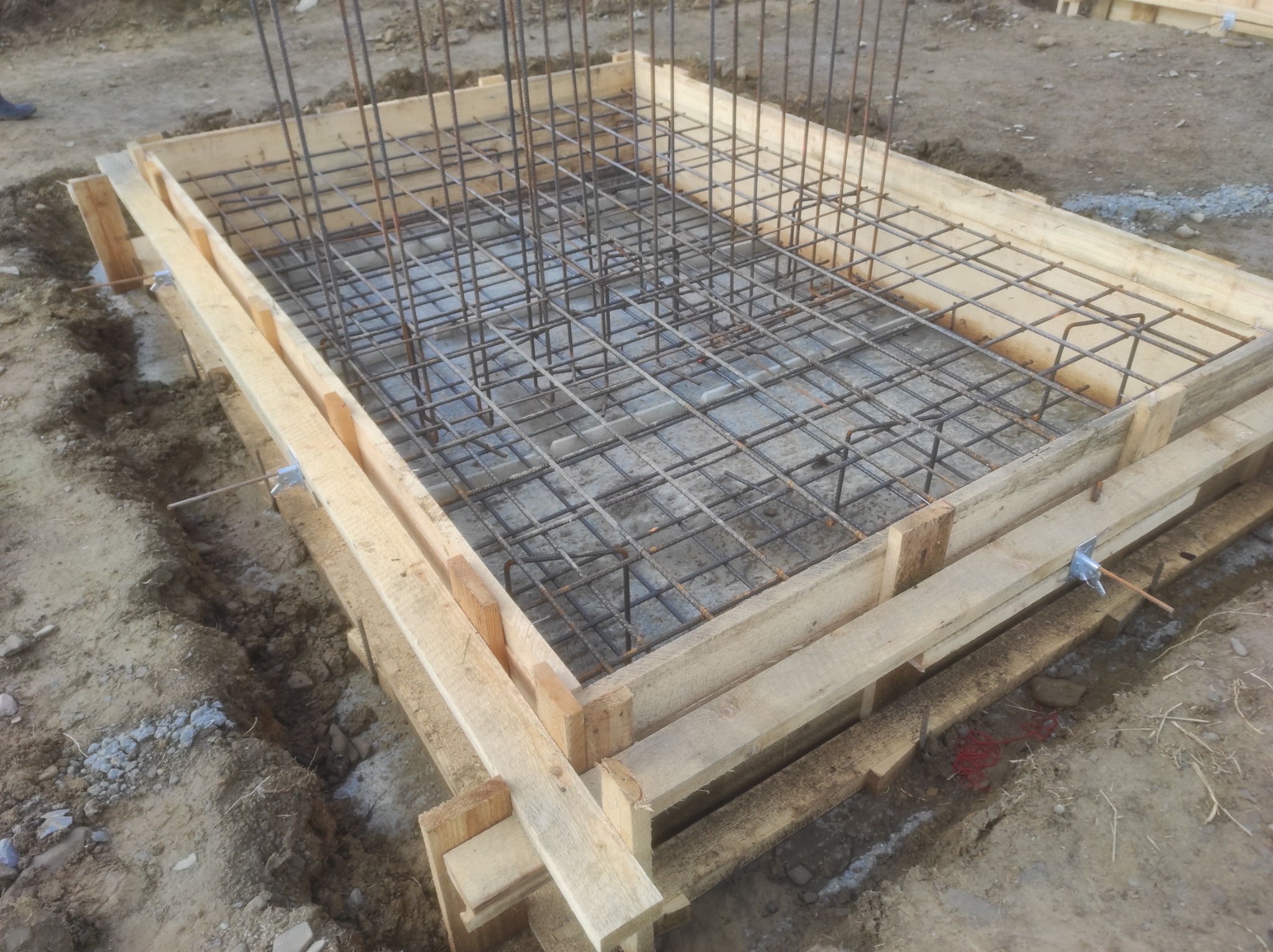 Szczegółowy widok szalunku pod fundament z ułożonym zbrojeniem, zabezpieczonym drewnianymi stemplami i metalowymi kotwami, otoczony ziemią budowlaną.