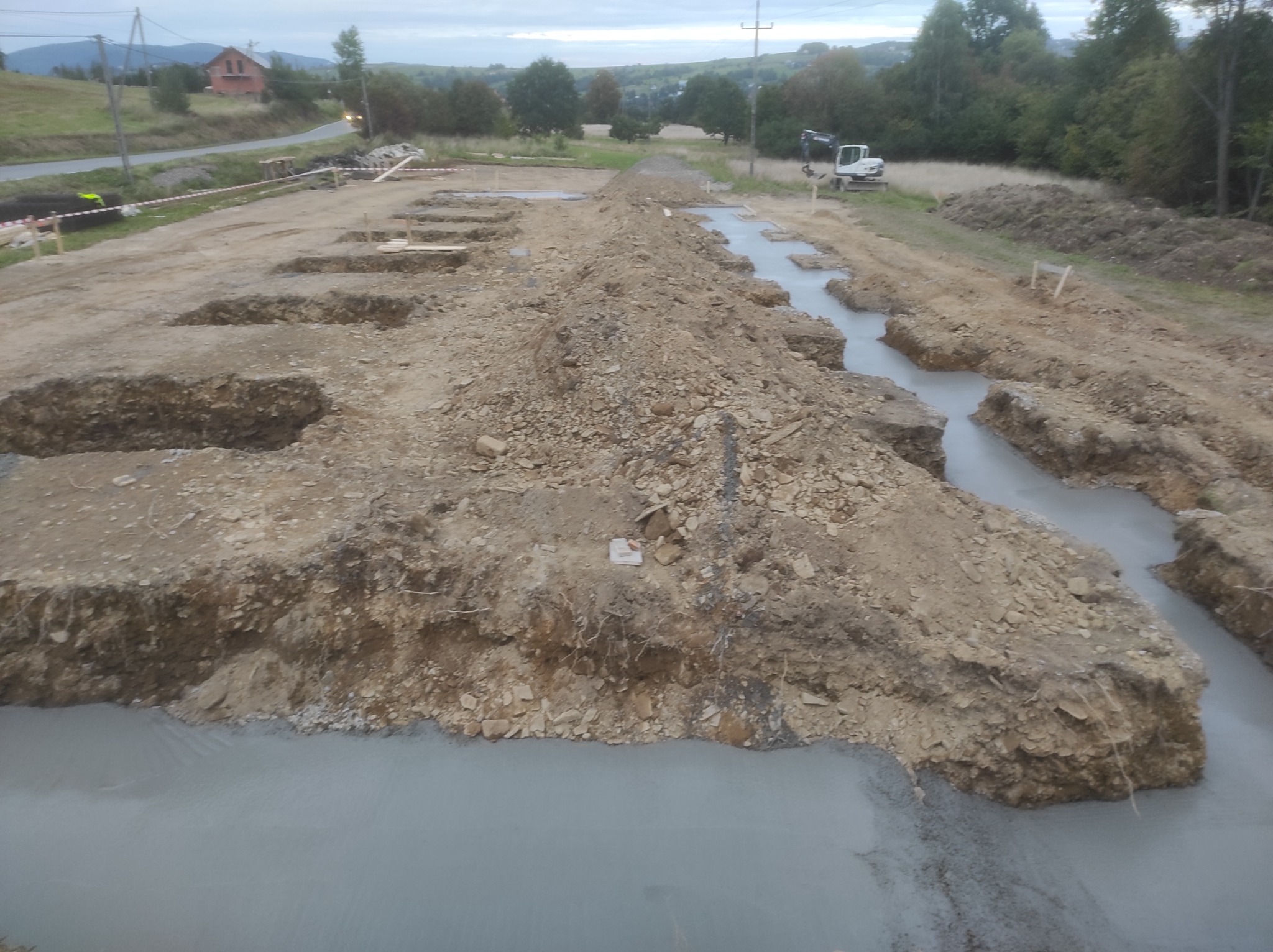 Wykopane rowy fundamentowe częściowo wypełnione betonem na placu budowy z widoczną koparką w tle, krajobraz pagórkowaty.