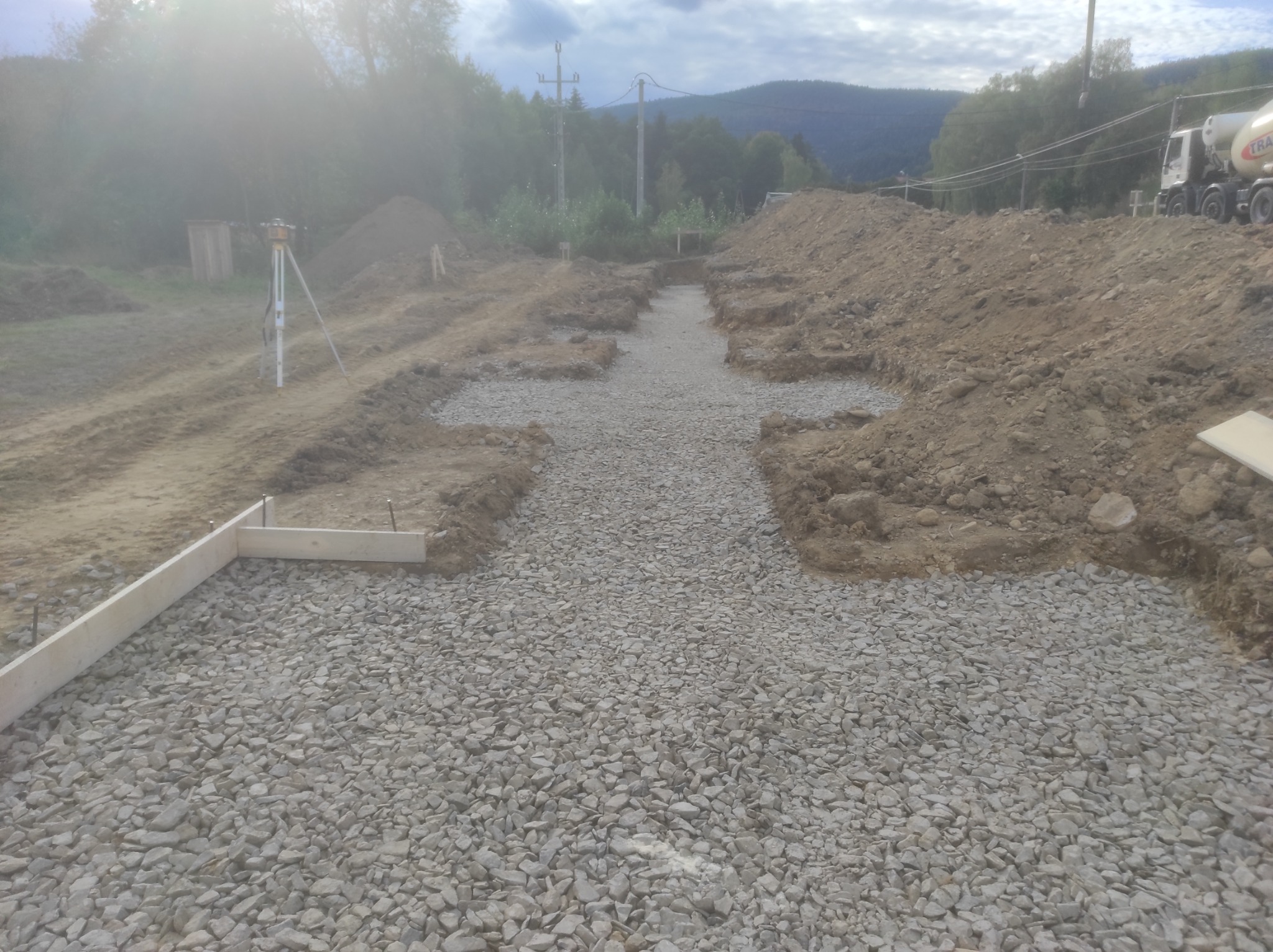 Przygotowanie terenu pod budowę: wykopane rowy zalane żwirem, widoczny niwelator laserowy, drewniane szalunki i betonowóz w tle.