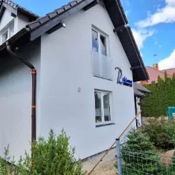 Odnowiona elewacja domu jednorodzinnego w Warczu, z nowymi oknami i szarym dachem, widok z poziomu ogrodu z zielenią. Na ścianie widoczne logo firmy.