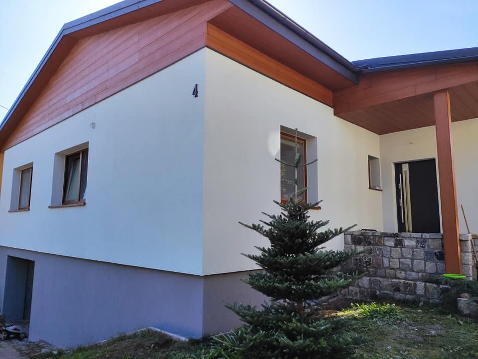 Elewacja domu jednorodzinnego z numerem 4 w Warczu, świeżo po tynkowaniu, z widocznym fragmentem poddasza wykończonego drewnem i kamiennym wejściem.