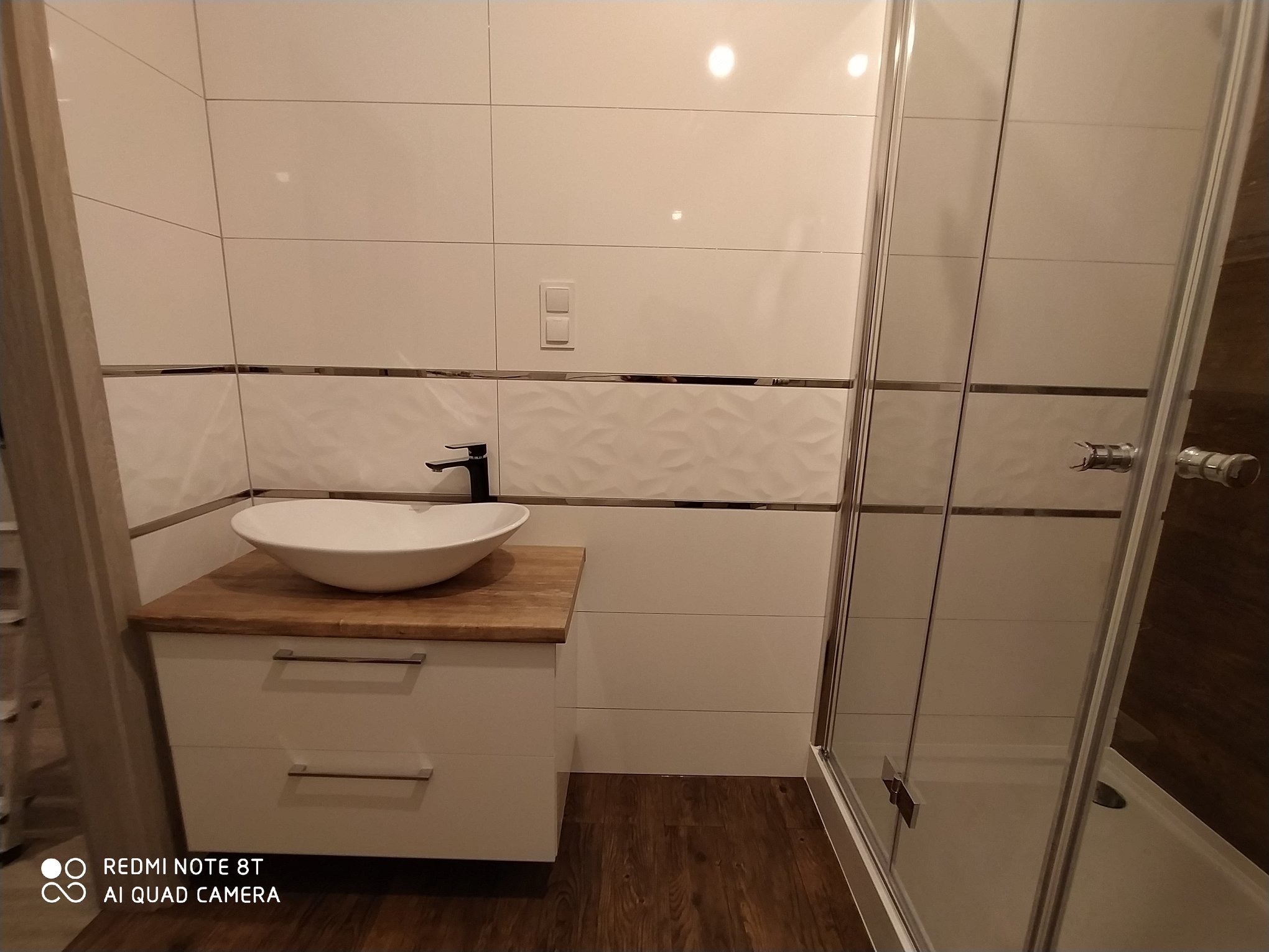 Nowoczesna łazienka z białą umywalką nablatową na drewnianym blacie, białe płytki z dekoracyjnym pasem i kabina prysznicowa ze srebrnymi ramami.