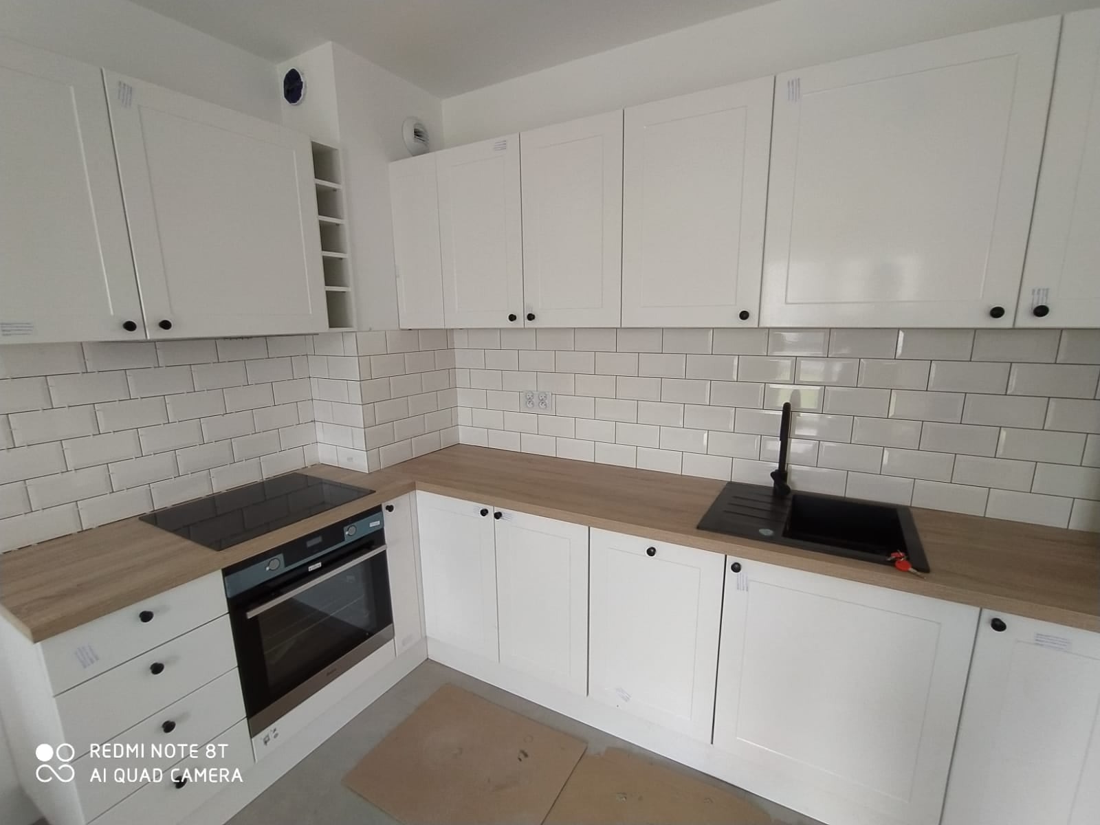 Nowoczesna, jasna kuchnia z białymi szafkami, drewnianym blatem i białymi płytkami metra na ścianie, widoczny czarny zlew i płyta indukcyjna.