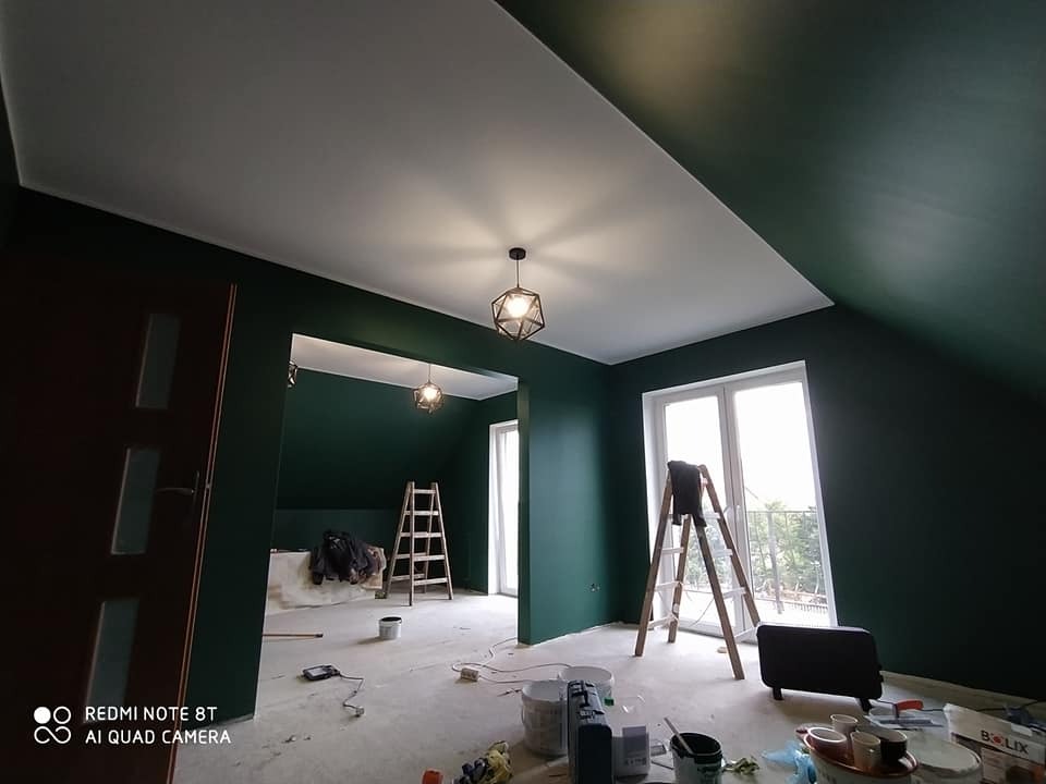 Pokój na poddaszu z pomalowanymi na ciemnozielono ścianami, biały sufit, drabiny, widoczne ślady prac malarskich na podłodze, nowoczesne lampy wiszące.