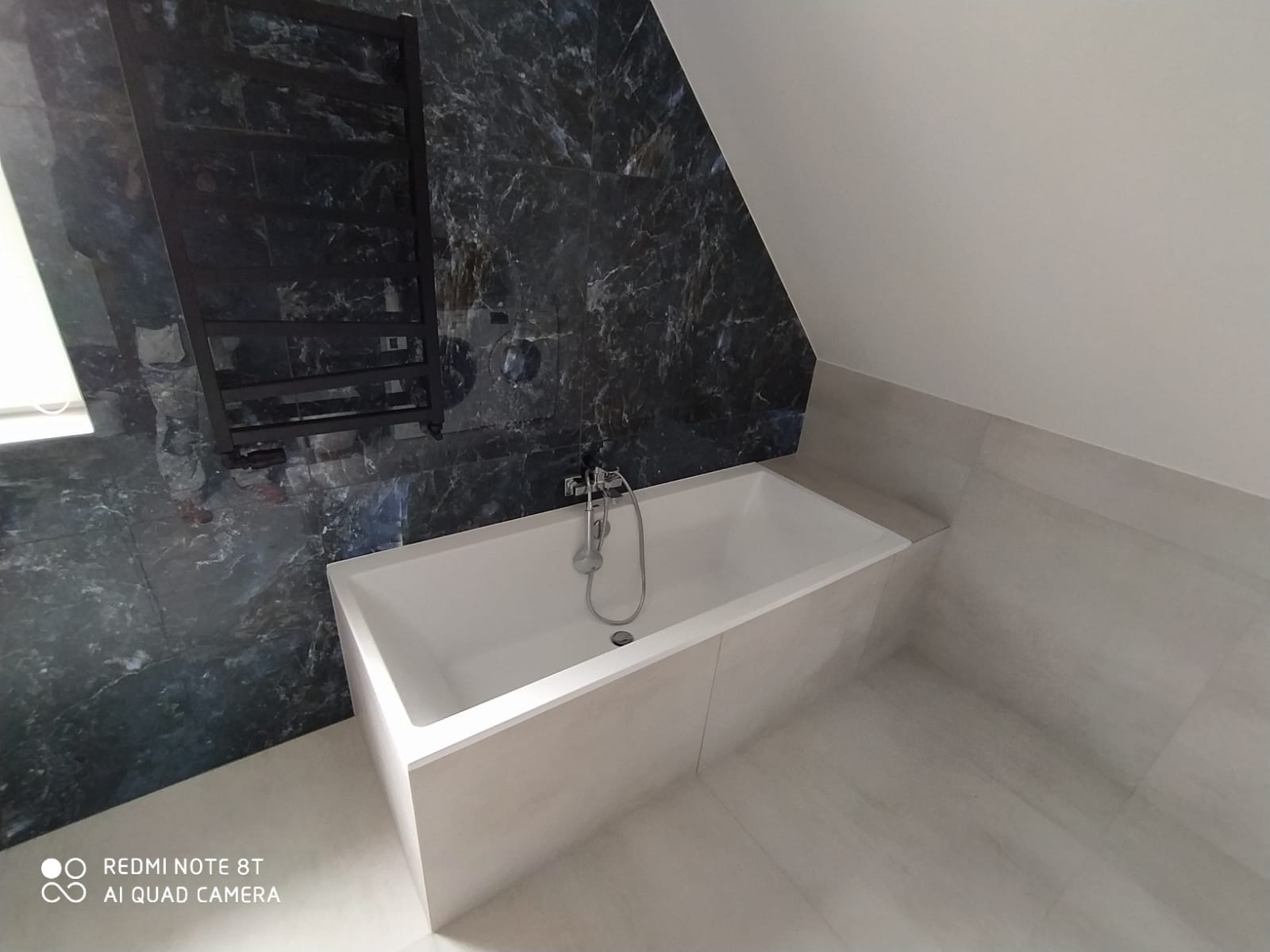 Wanna prostokątna obudowana płytkami w łazience na poddaszu, z baterią chromowaną i grzejnikiem drabinkowym na ścianie wyłożonej marmurkowymi płytkami.