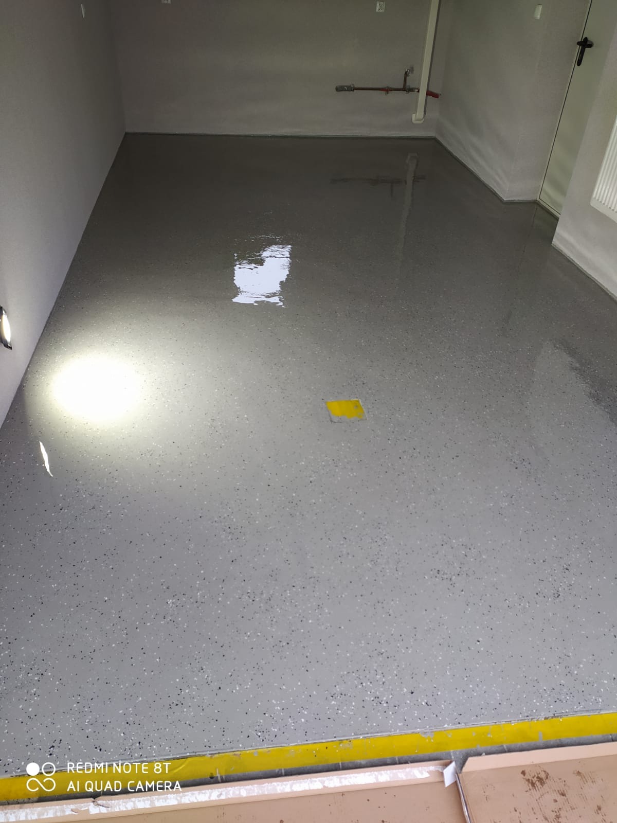 Świeżo wylana, szara, żywiczna posadzka z drobnymi, czarnymi i białymi elementami, z połyskiem, w pustym pomieszczeniu z widocznymi instalacjami na ścianie i żółtą taśmą ochronną przy podłodze.