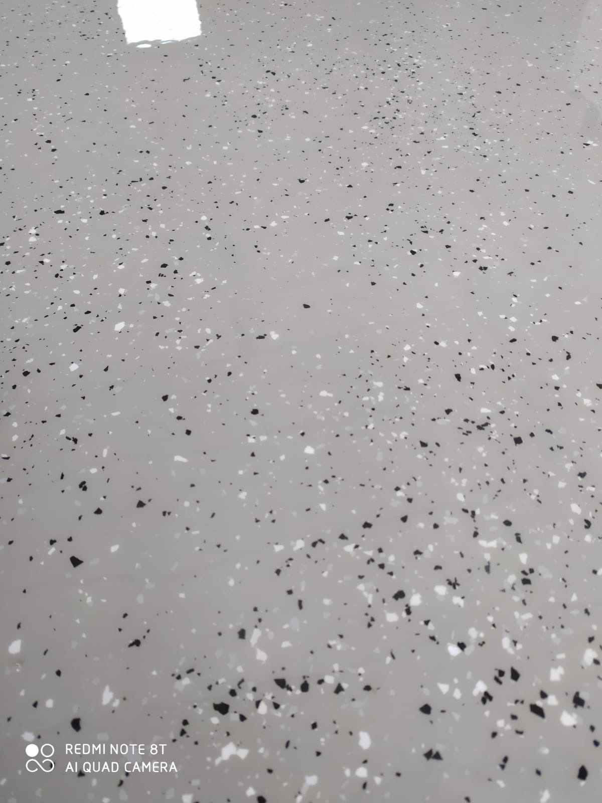 Błyszcząca, jasnoszara posadzka żywiczna z czarnymi i białymi drobinami, widoczne odbicie światła, ujęcie z góry.