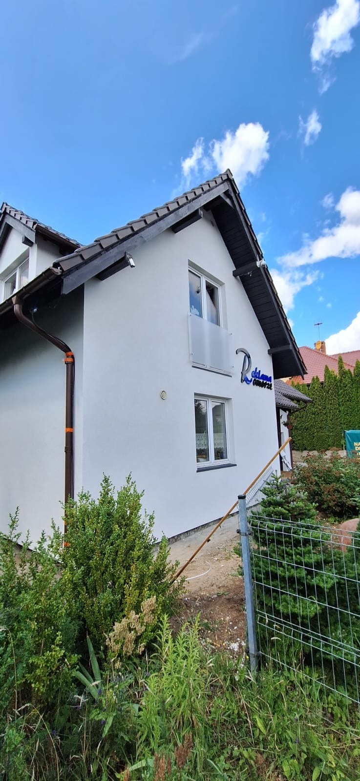 Odnowiona elewacja domu jednorodzinnego w Warczu, z nowymi oknami i szarym dachem, widok z poziomu ogrodu z zielenią. Na ścianie widoczne logo firmy.