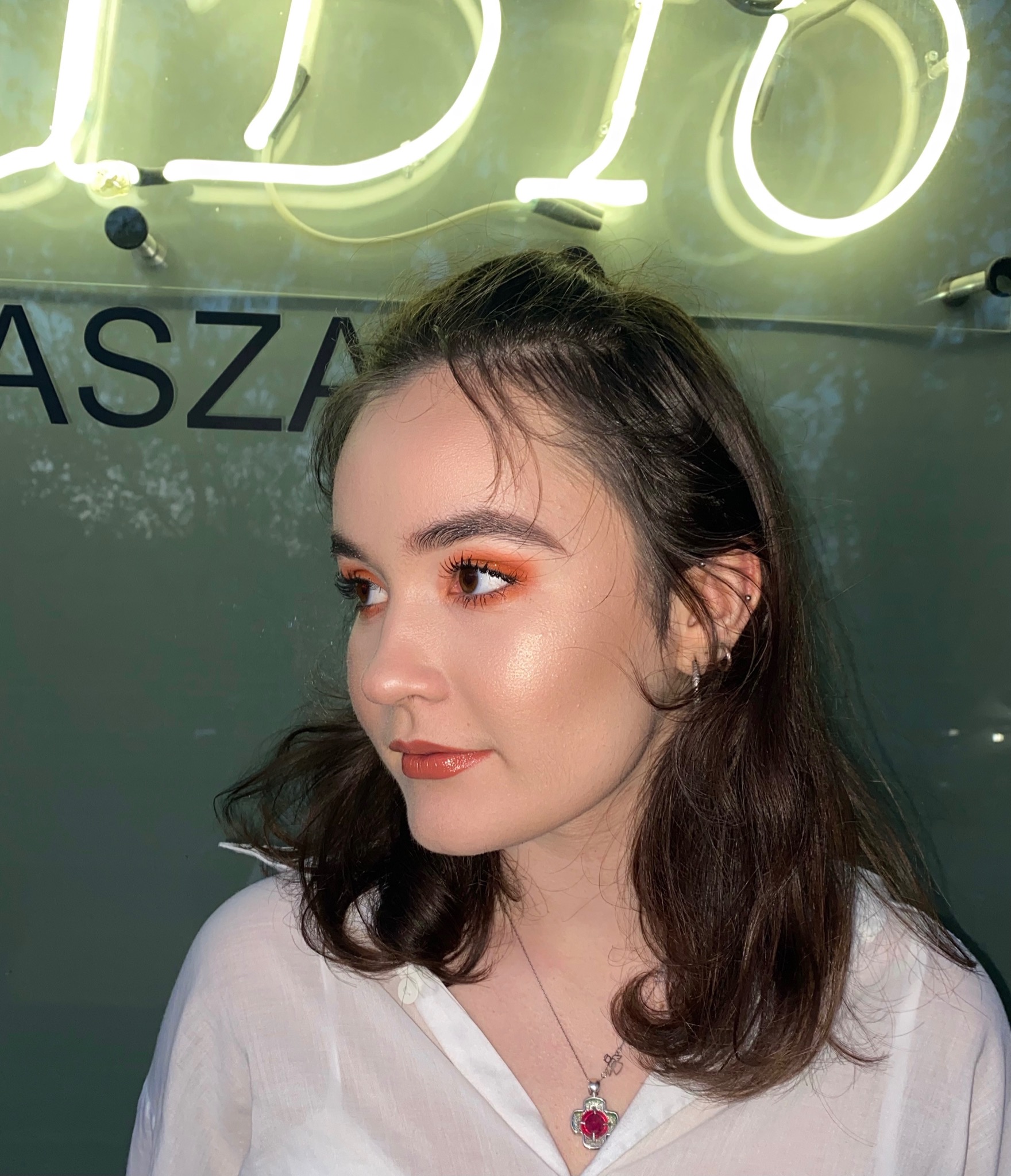 Portret kobiety z pomarańczowym makijażem oczu i błyszczącą cerą, na tle neonowego napisu.