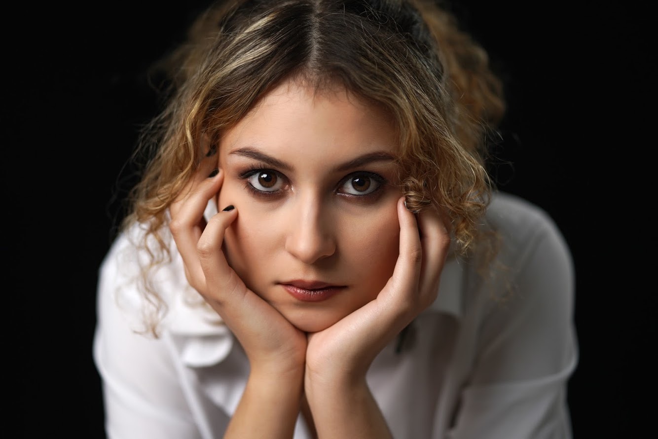 Portret kobiety z delikatnym makijażem oczu i ust, opierającej twarz na dłoniach z czarnym manicure na tle czerni.