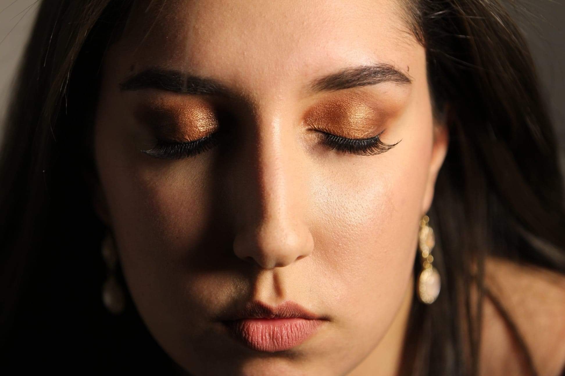 Portret kobiety z zamkniętymi oczami, prezentujący złoty, błyszczący cień do powiek i sztuczne rzęsy, z widocznymi delikatnymi kolczykami w uszach.