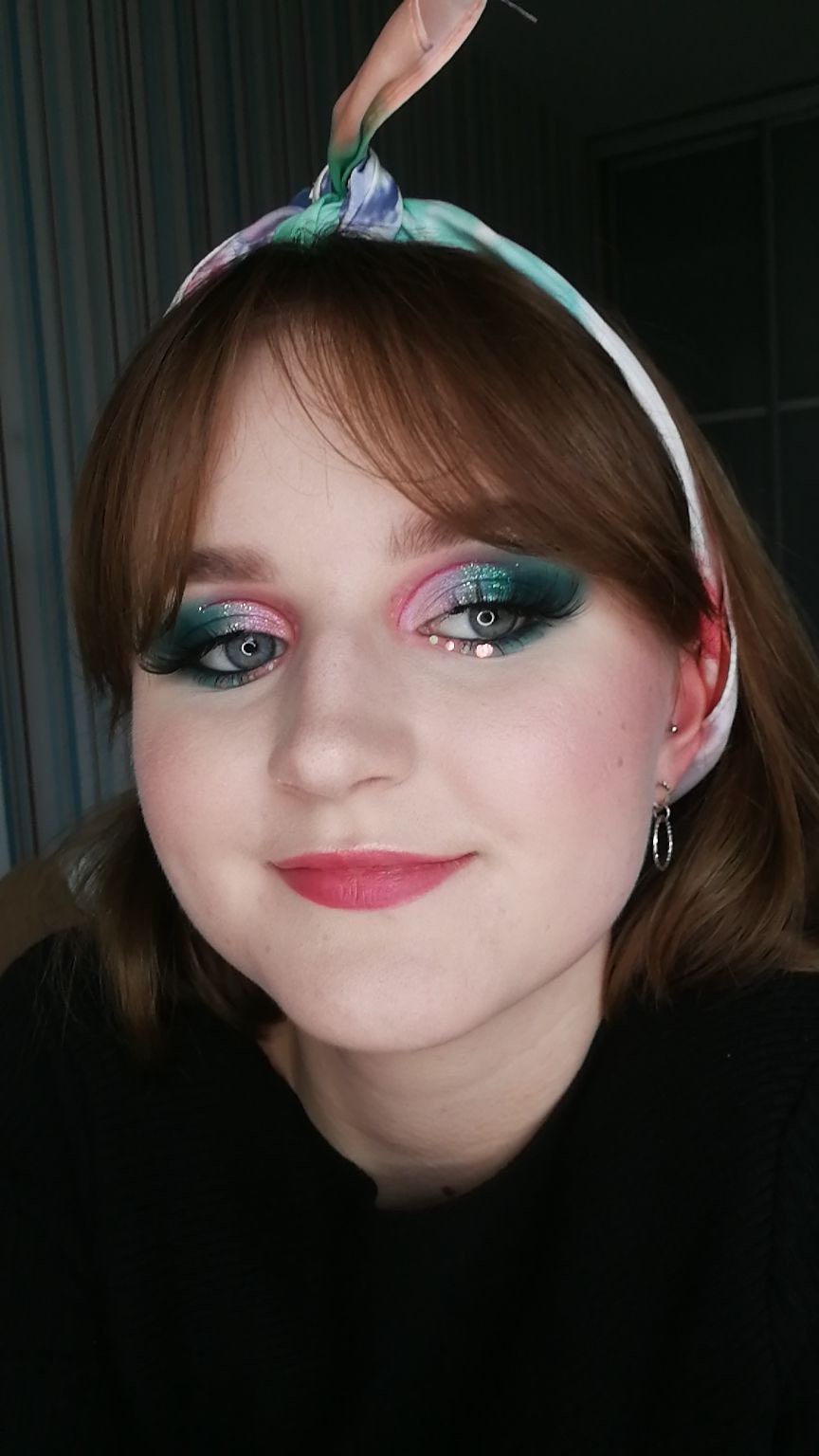 Portret kobiety z artystycznym makijażem oczu w odcieniach różu i zieleni, ozdobionym małymi kryształkami, oraz z różową pomadką i opaską na włosach.