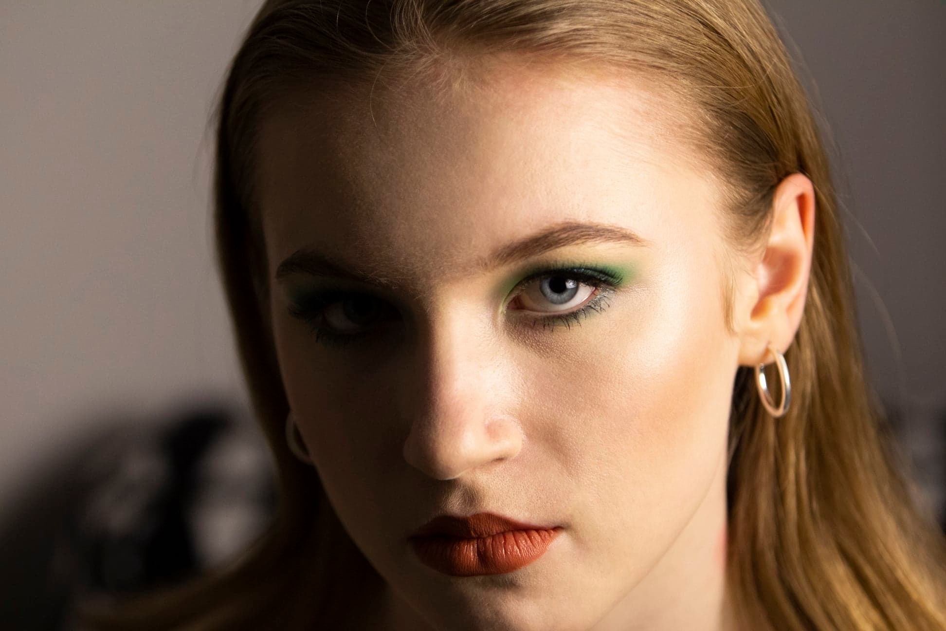 Portret kobiety z intensywnym zielonym cieniem na powiekach i pomarańczową szminką, podkreślone kości policzkowe, srebrne kolczyki koła.