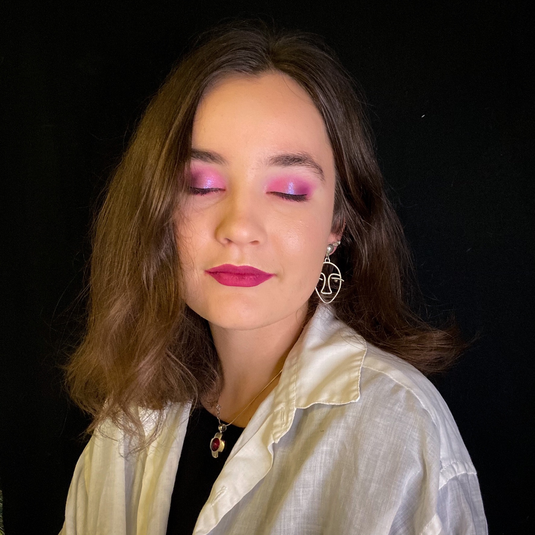Portret kobiety z zamkniętymi oczami i intensywnym, różowym makijażem oczu i ust; srebrne kolczyki z motywem twarzy, delikatny łańcuszek z czerwonym kamieniem na czarnym tle.