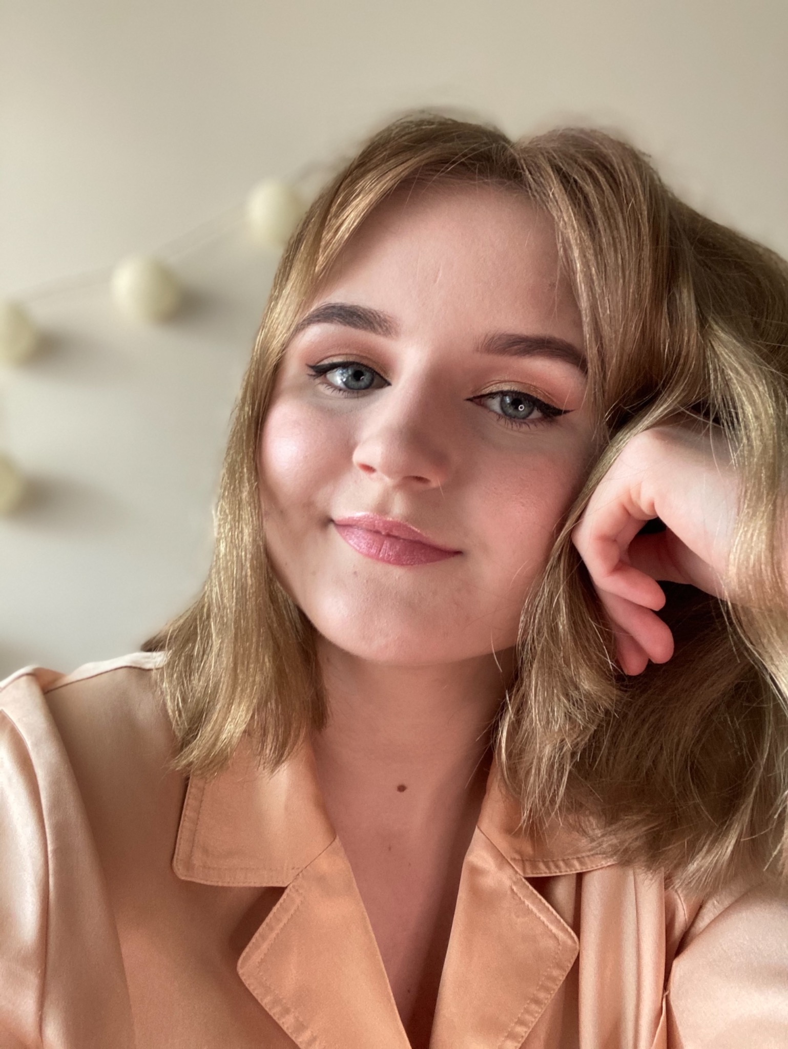 Portret uśmiechniętej blondynki z delikatnym makijażem oczu i ust, opierającej głowę na dłoni, w satynowej koszuli.