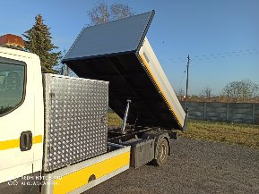 Wywrócony kontener na samochodzie dostawczym z widocznym mechanizmem hydraulicznym podnoszenia, żółte elementy odblaskowe, błękitne niebo w tle.