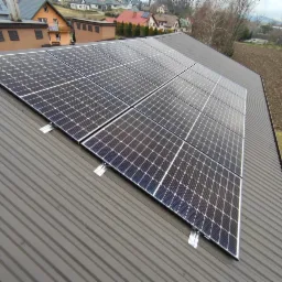 Panel słoneczny na brązowym dachu blaszanym, widok z góry, w tle zabudowa jednorodzinna i pole uprawne.