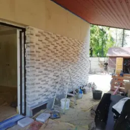 Układanie dekoracyjnej okładziny imitującej cegłę na elewacji budynku, widoczna drabina, wiadro z zaprawą, narzędzia i zabezpieczenia podłoża.